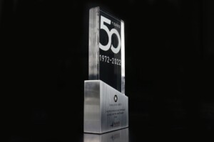 A transparent glass trophy commemorating 50 years, from 1972 to 2022, displayed against a black background. the recognition award has an inscription and logos.