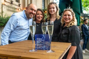 The Thinkbox TV Awards held at One Marylebone Road, 12th July 2023Photography by Fergus Burnett Four happy adults stand behind a wooden table with three glasses of champagne and a "thinkbox tv planning recognition awards 2023" trophy, celebrating outdoors.
